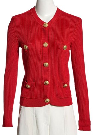 Cardigan en maille côtelée rouge avec manches longues, boutons dorés sur le devant et quatre petites poches avant, porté avec un pantalon blanc.