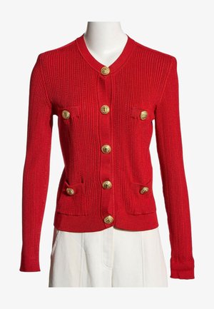 Cardigan en maille côtelée rouge avec manches longues, boutons dorés sur le devant et quatre petites poches avant, porté avec un pantalon blanc.