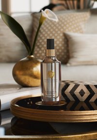 Glasparfümflasche mit einem goldenen Deckel und Emblem, beschriftet mit "White Basil", auf einem runden goldenen Tablett vor einem gemusterten Hintergrund platziert.