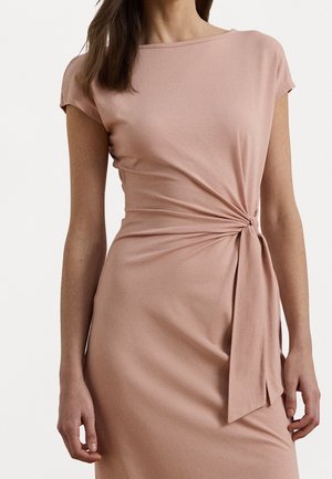 Femme portant une robe rose pâle à manches courtes avec un détail de nœud sur le côté qui crée un effet froncé au niveau de la taille.