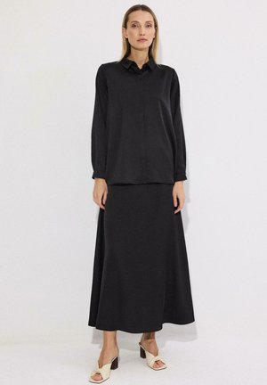 Γυναίκα με μακριά μαύρη φούστα και άνετο μαύρο πουκάμισο με γιακά, στέκεται με χαλαρά χέρια, λευκό φόντο, μπεζ πέδιλα με τακούνια.