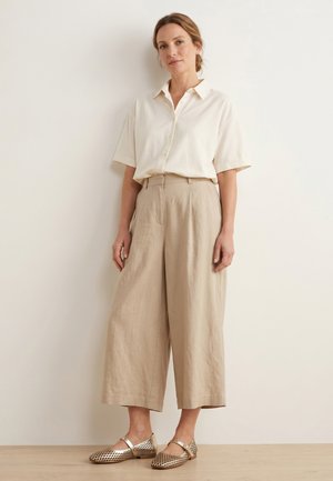 Kobieta opierająca się o ścianę, ubrana w kremową, krótką koszulę na guziki, beżowe szerokie spodnie 7/8 oraz metaliczne, tkane baleriny.