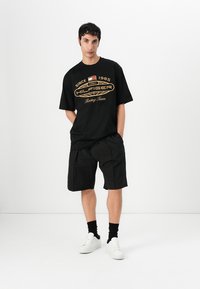 Svart T-skjorte med gull tekst, "HILFIGER Racing Team," kombinert med svarte shorts. Hvite joggesko og svarte sokker fullfører antrekket.