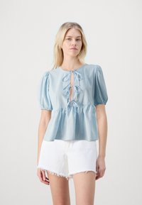 Blusa di cotone azzurro chiaro con maniche a sbuffo, un'apertura a forma di chiave e dettagli con cordoncino sullo scollo, abbinata a pantaloni corti bianchi con un look consumato.