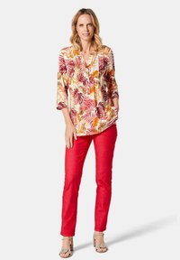 Blumenhemd mit orangefarbenen und maronifarbenen Blättermustern, kombiniert mit schmalen roten Hosen und hellgrauen Sandalen, stehend vor einem weißen Hintergrund.