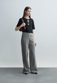 Mujer de pie vistiendo una blusa negra de manga corta con escote de ojal, pantalones grises de pierna ancha, zapatos negros y llevando un bolso de hombro marrón oscuro.