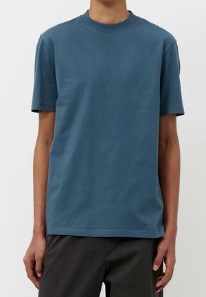 T-shirt bleu à manches courtes et col rond, porté par une personne avec les bras et le torse visibles, assorti à un pantalon foncé sur un fond uni.
