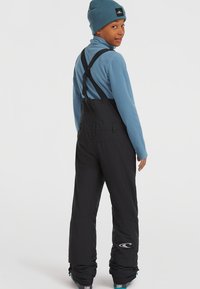 Schwarze Skihose mit verstellbaren Trägern, kombiniert mit einem hellblauen Fleece-Oberteil und einer blauen Strickmütze. Mit Logodetail am Hosenbein.
