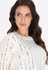 Weißer Strickpullover mit vertikalen silbernen und schwarzen Paillettenstreifen. Verfügt über einen runden Ausschnitt und eine weiche Textur. Akzentuiert mit subtiler Ausstrahlung.