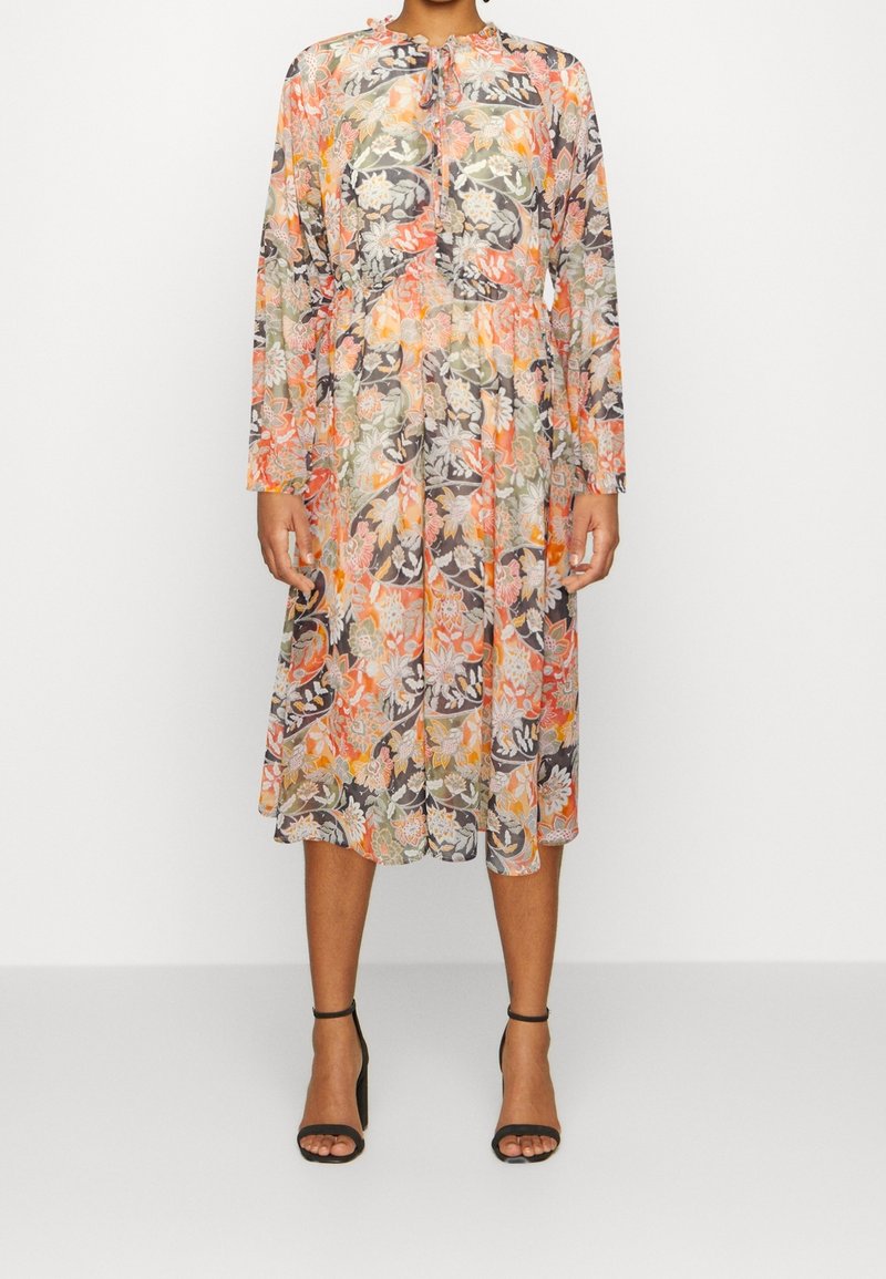 Femme portant une robe à manches longues, mi-longue, à motifs floraux orange, blancs et verts, associée à des escarpins noirs à bride à la cheville.
