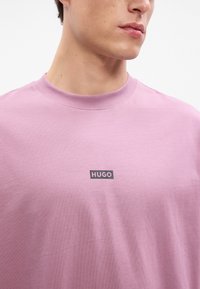 Krátky rukáv, tričko s okrúhlym výstrihom z mäkkej ružovej bavlny s čiernou etiketou s logom "HUGO" umiestnenou na strede hrude. Hladká textúra.