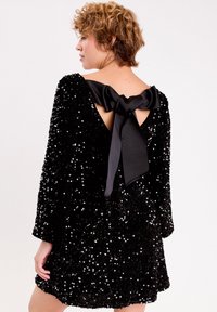 Robe en sequins noirs avec des manches longues et un dos nu, ornée d'un large nœud en satin noir au niveau du cou et d'une silhouette évasée.