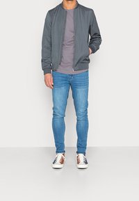 Chaqueta bomber masculina de tela gris oscuro, combinada con una camiseta gris clara y jeans ajustados azules. El calzado presenta una base blanca con acentos azules y marrones.