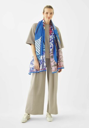 Vrouw draagt beige wijde broekspijpen en een oversized T-shirt, witte sneakers, en een lange, kleurrijk patroon sjaal om haar nek gedrapeerd.