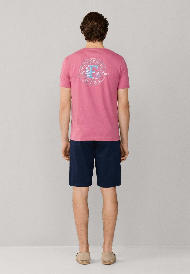 Uomo che indossa una maglietta rosa a maniche corte con logo circolare "Faconnable" sul retro, pantaloncini blu navy e scarpe slip-on beige, in piedi contro un muro grigio.