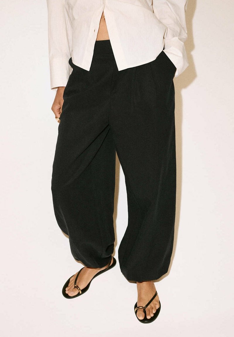 Personne portant un pantalon noir ample, une chemise blanche boutonnée partiellement rentrée, et des sandales noires, debout devant un fond uni.