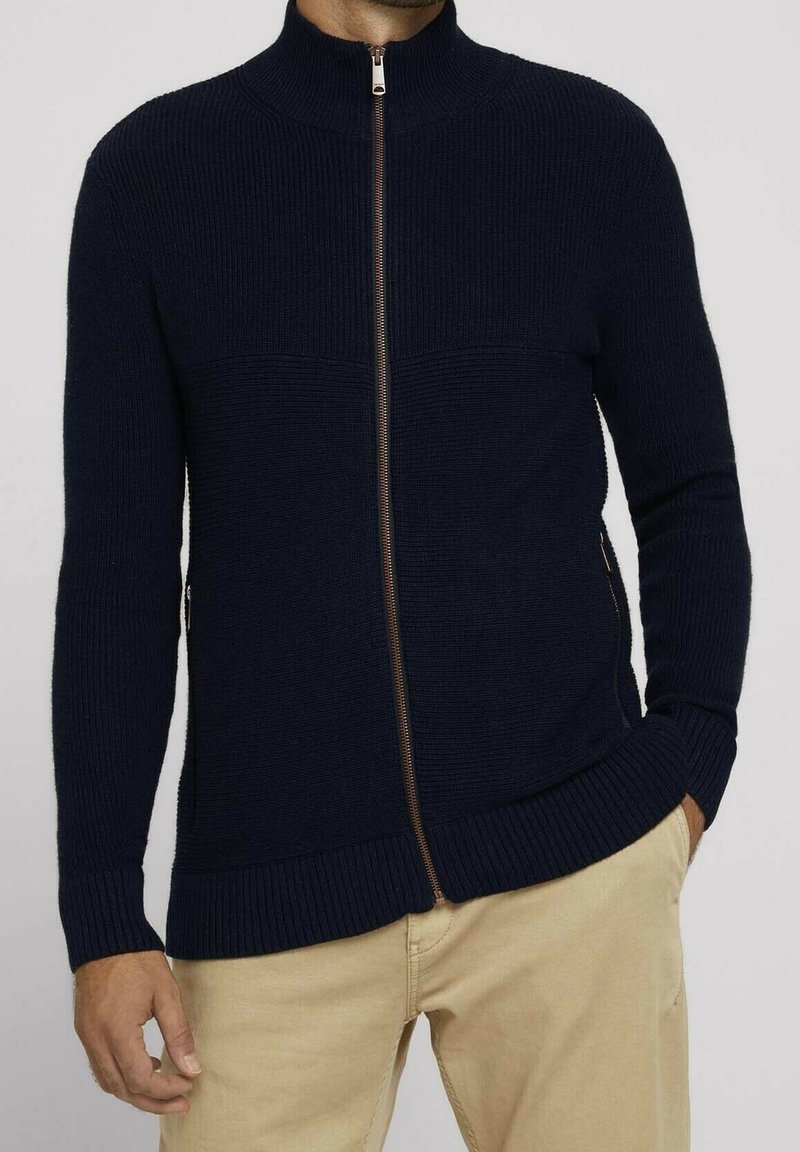 Pull zip noir avec col haut, texture côtelée et fermeture éclair contrastante. Il dispose de poches latérales et d'un design ajusté.