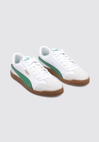 Zapatillas blancas con detalles en verde, puntera de ante y suela de goma marrón. Presenta cordones blancos y un logotipo en la lengüeta y el costado.