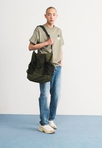 Sac en toile vert avec des accents en maille noire, grande forme et doubles poignées. Porté par une personne en chemise grise, jeans bleus et baskets blanches.