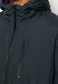 Giacca con cappuccio di un grigio scuro, realizzata in materiale liscio e resistente all'acqua; presenta una chiusura centrale con zip e una tasca sul petto con zip.