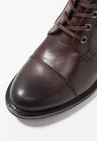 Bota de cuero marrón con puntera redonda, costuras detalladas, ojales de metal y cordones marrones. Textura suave y acabado pulido.