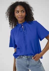Haut bleu texturé à manches courtes avec un col volanté, détail de nœud et patte de boutonnage, associé à un jean en denim bleu clair.