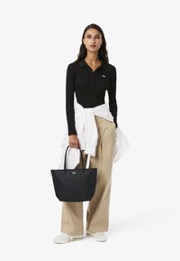Svart långärmad polo med en grön logotyp, beige vida byxor, vit wrap och en stor svart tote-väska med två handtag.