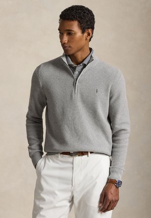 Mann iført lys grå teksturert genser med kvart-zip over svart-hvitt stripet polo, hvite bukser, brun belte og klokke med brun skinnreim.