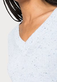 Ljusblått stickat tröja med V-hals, med ett texturerat mönster och fläckar av mörkblått och svart genom tyget.