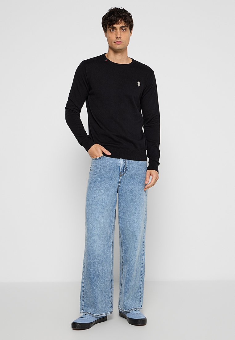Schwarzer langärmeliger Strickpullover mit rundem Ausschnitt, kombiniert mit hellblauen weit geschnittenen Jeans und schwarzen Schuhen. Schlichte Stickdetails auf dem Pullover.