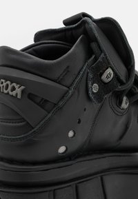 Black leather shoe with rounded shape, featuring Velcro strap, metallic hardware accents, and perforated detailing for breathability.