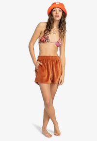 Top bikini con motivo floreale e spalline sottili, abbinato a pantaloni corti arancioni, morbidi e texturizzati, con vita elastica e tasche laterali.