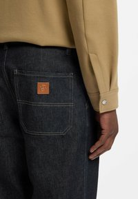 Camicia beige a maniche lunghe con dettaglio sul polsino abbinata a pantaloni in denim blu scuro con etichetta in pelle marrone sulla tasca posteriore.