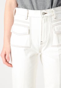 Vita denimplagg med synliga sömmar, två stora framfickor och metallknappstängning. Formade för att passa vid midjan.