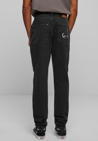 Juodi džinsai su tiesiu šoniniu kirpimu, užpakalinėmis kišenėmis, siūtu logotipo detaliu ir odiniu lopu ant juosmens.