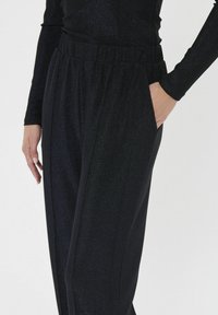 Crne hlače s elastikom u struku i sjajnom teksturom. Imaju bočne džepove i širok kroj, koji hvata svjetlost za suptilan efekt sjaja.