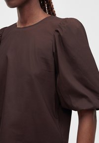 Gros plan d'une personne portant un chemisier marron foncé avec des manches courtes bouffantes et un col rond.