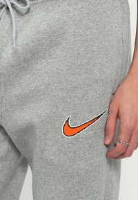 Pelēki treniņbikses ar strukturētu audumu, elastīgu jostasvietu un izteiksmīgu oranžu Nike logotipu uz kreisā augšstilba.