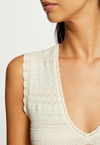 Strickoberteil in cremefarbener Ausführung mit tiefem V-Ausschnitt, gewellten Ärmeln und kunstvollen strukturierten Mustern auf dem Stoff.