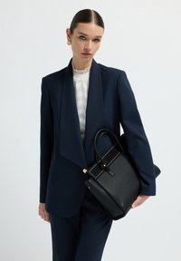 Žena v námořnickém obleku držící černou koženou kabelku, nosící zlaté náušnice a bílý strukturovaný top s volánovým límcem.