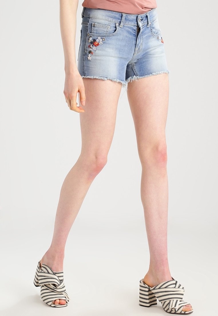 Femme portant un short en jean bleu clair brodé et des sandales à talons rayées noir et blanc, debout avec les jambes croisées.