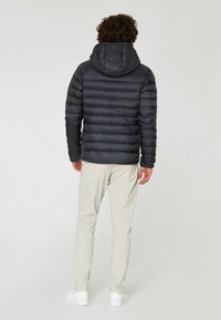 Vadderad svart jacka med huva, med horisontell quiltad design. Slät textur och minimal varumärkning på baksidan. Kombinerad med ljusa byxor.