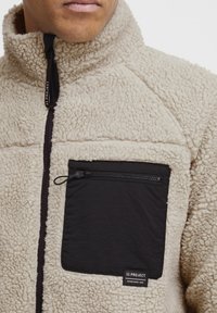 Fleece jacket in beige with a high collar, featuring a black zippered pocket and contrasting stitching. Textured fabric adds warmth.