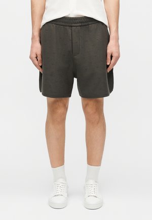 Muž nosiaci tmavosivé šortky s jemným reliéfnym vzorom, biele ponožky a biele tenisky, stojaci pred jednofarebným pozadím.