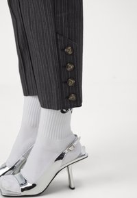 Pantaloni scuri a righe sottili con una tasca laterale e quattro bottoni all'orlo. Scarpe con tacco alto argentate e calzini bianchi completano l'outfit.
