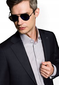 Czarna marynarka na tle lekko szarej i białej w paski koszuli. Szare okulary przeciwsłoneczne z ciemnymi szkłami, wypolerowane detale i zapięty kołnierz.