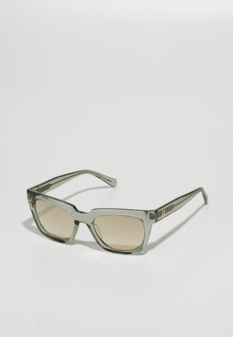 Ochelari de soare pătrați cu un cadru din plastic verde translucid, lentile cu un gradient de culoare maro deschis și branduire subtilă pe braț.