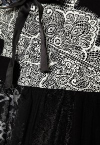 Schwarzes Kleid mit einem strukturierten Spitzenoberteil in Weiß mit komplizierten Mustern, einem schwarzen Band am Hals und einem fließenden schwarzen Rock.