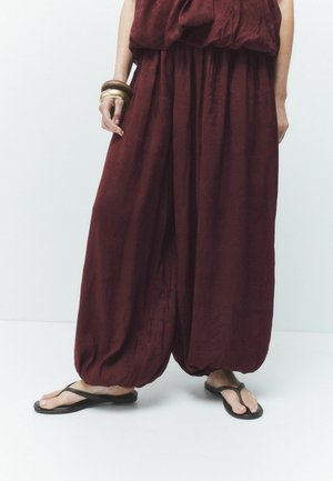 Person trägt lockere bordeauxrote Hose und passendes Oberteil, schwarze Flip-Flops und klobige Armbänder am linken Handgelenk, steht auf weißem Boden.