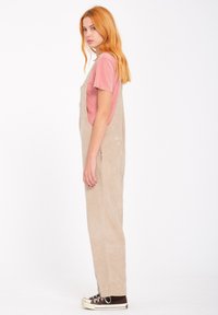 Beige kordfløjsoverall har en afslappet pasform, sidelommer og justerbare stropper, stylet over en lyserød T-shirt og brune sneakers.